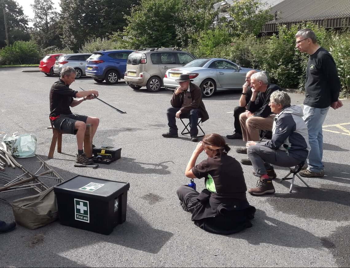 A group of volunteers being taught how to sharpen a scythe blade